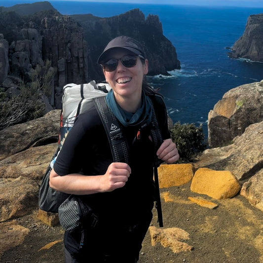 Person with a backpack standing on a rocky cliff overlooking the ocean. Location is Three Capes in Tasmania. The person is wearing Bluey Merino hike t-shirt in black © Bluey Merino Tasmania