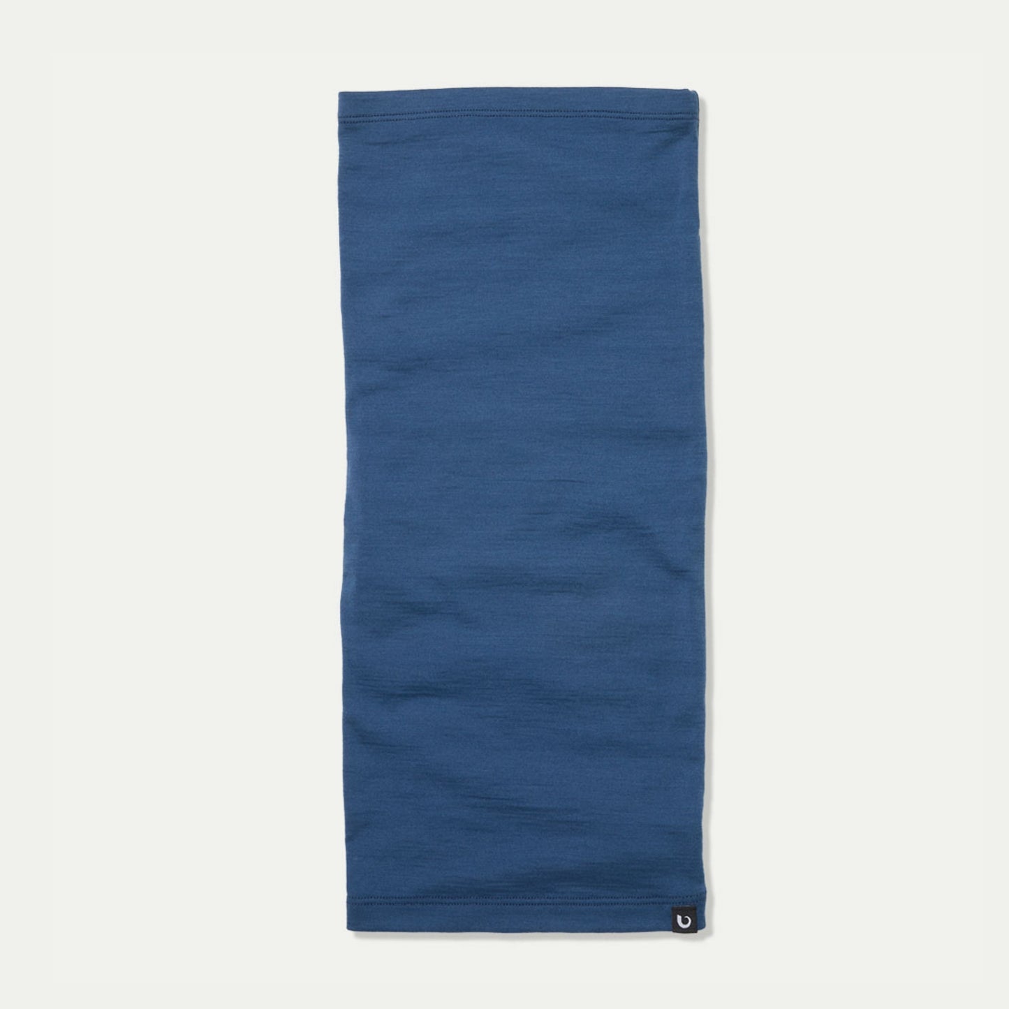 A solid blue French navy neck gaiter laid flat on a light off-white background, featuring a small Bluey Merino black logo patch at the bottom right hem. © Bluey Merino Tasmania