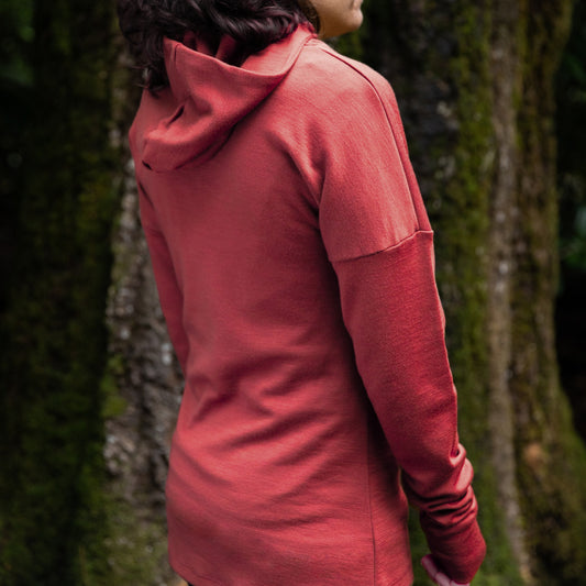 Women is wearing the Merino Tullah Hoodie - Rose Robin by © Bluey Merino Tasmania. She is standing in front of a large tree trunk turning her back to us.