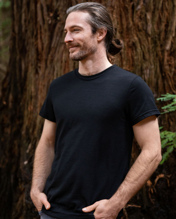 Steve is wearing Men's Tasmanian Merino 180 Emrik Classic Crew Neck T-Shirt in Currawong (black). He is standing in front of a large tree trunk in Hollybank reserve park in Northern Tasmania. Photography by Melanie Kate Creative -  © Bluey Merino