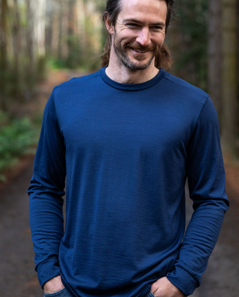 Men's Tasmanian Merino 180 Orson Long Sleeve Crew Neck Top . The top has wrist cuffs and a fine stitching line across the top shoulders. The crew neck line is classic and hugs the neck nicely - Steve is smiling and is  relaxed with his hands in his jeans pockets. He is standing in the middle of Hollybank reserve (Tasmania) amongst trees. Photography by Melanie Kate Creative -  © Bluey Merino