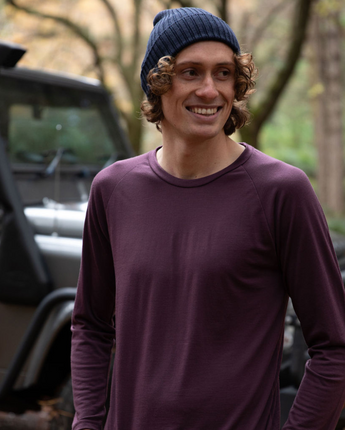 Hamish is wearing the Men’s Tasmanian Merino 180 Raglan Long Sleeve Top in Dark Bridestowe with the Fisherman Beanie in Navy. He is smiling, relaxed with hands in pockets standing in front of BLU the Defender 110 - Photography by Melanie Kate Creative -  © Bluey Merino
