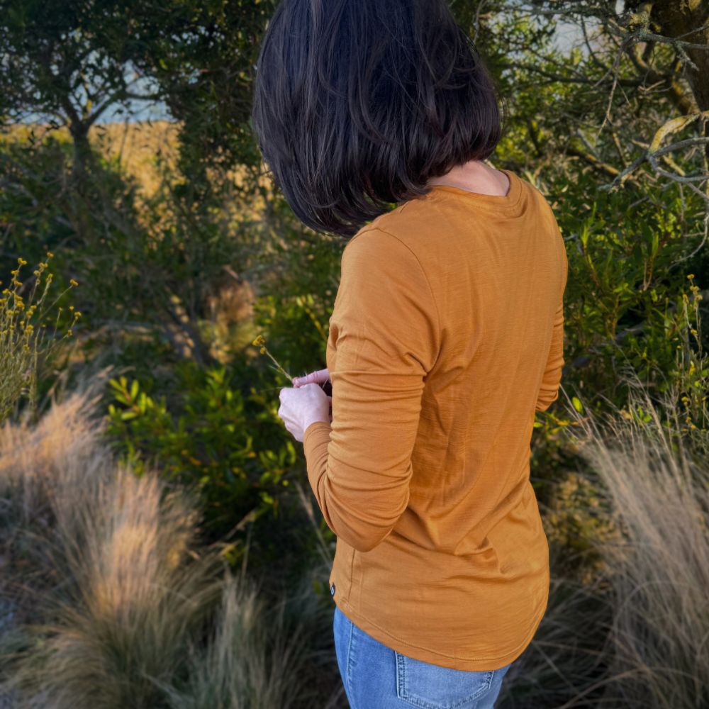 Person wearing a mustard yellow - Billy Button long-sleeve Merino shirt standing in a natural setting with trees, grass and billy buttons flowers © Bluey Merino Tasmania