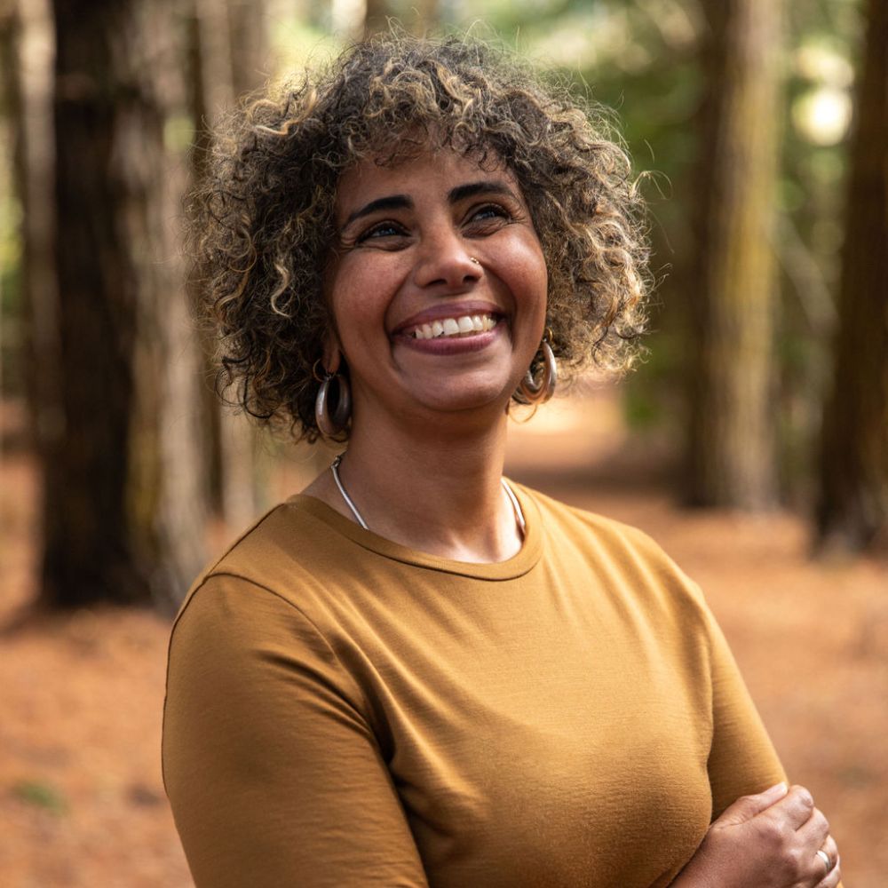 Woman with curly hair smiling in a forest setting. She is wearing a long sleeve Merino top in the colour Kunanyi - Bronze © Bluey 
