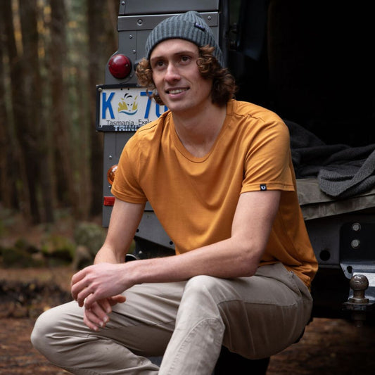 Man sitting at the back of a BLU the Defender 110 in Tasmanian wilderness. He is wearing a bring mustard yellow Merino t-shirt and a grey fisherman knit Merino beanie. © Bluey Merino Tasmania