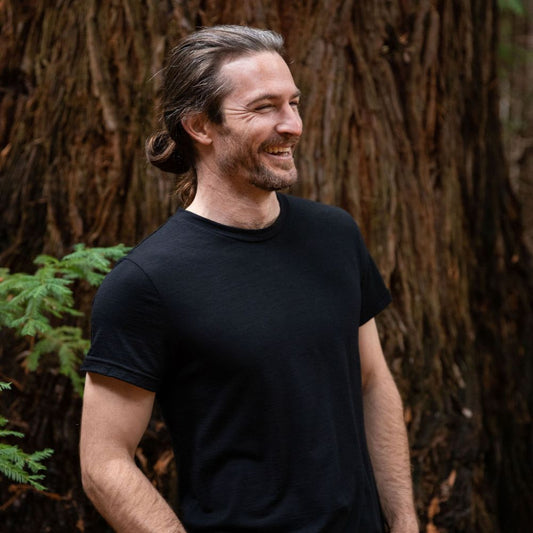 Laughing man with a beard and long hair standing in front of a large tree. He is wearing a black Merino t-shirt © Bluey Merino Tasmania