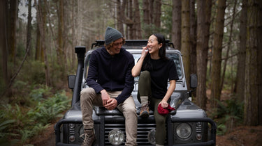 Two people laughing wearing Bluey Merino gear sitting on BLU the Classic Land Rover Defender 110. The scene is located in Tasmania's wilderness. Image © Bluey Merino Tasmania
