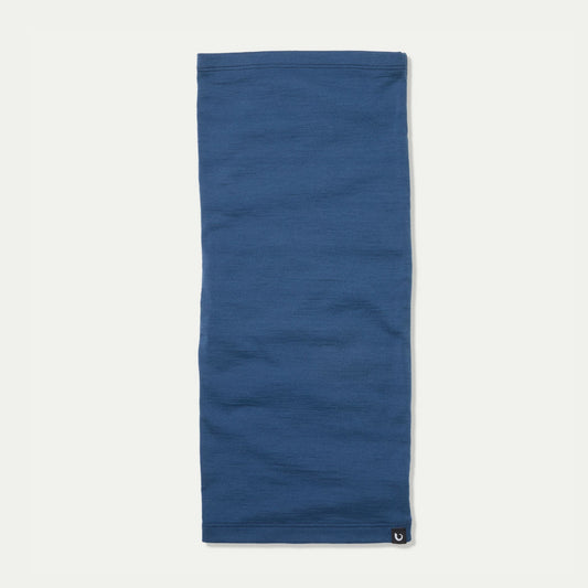 A solid blue French navy neck gaiter laid flat on a light off-white background, featuring a small Bluey Merino black logo patch at the bottom right hem. © Bluey Merino Tasmania
