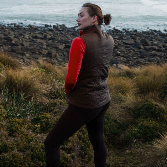 Person standing on a rocky coastal path wearing a brown Merino Gilet vest over a red shirt. © Bluey Merino Tasmania