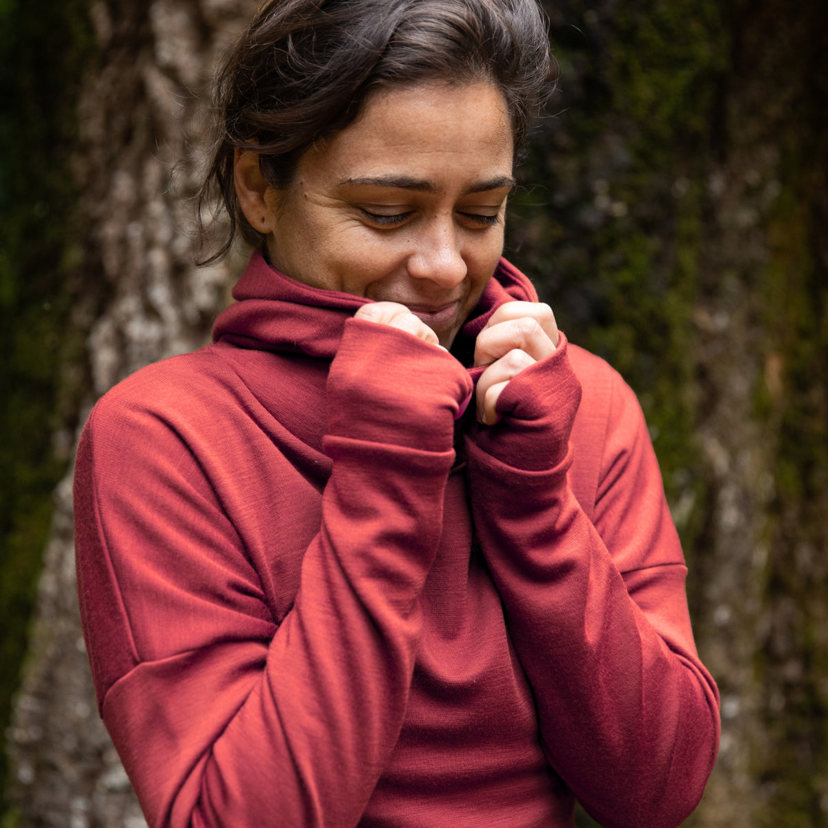 Women is wearing the Merino Nora Hoodie in colour Warm Spice Rose. She is snug and standing in front of a large tree trunk. ©Bluey Merino Tasmania