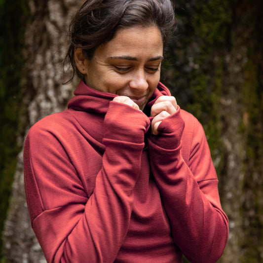 Women is wearing the Merino Nora Hoodie in colour Warm Spice Rose. She is snug and standing in front of a large tree trunk. ©Bluey Merino Tasmania